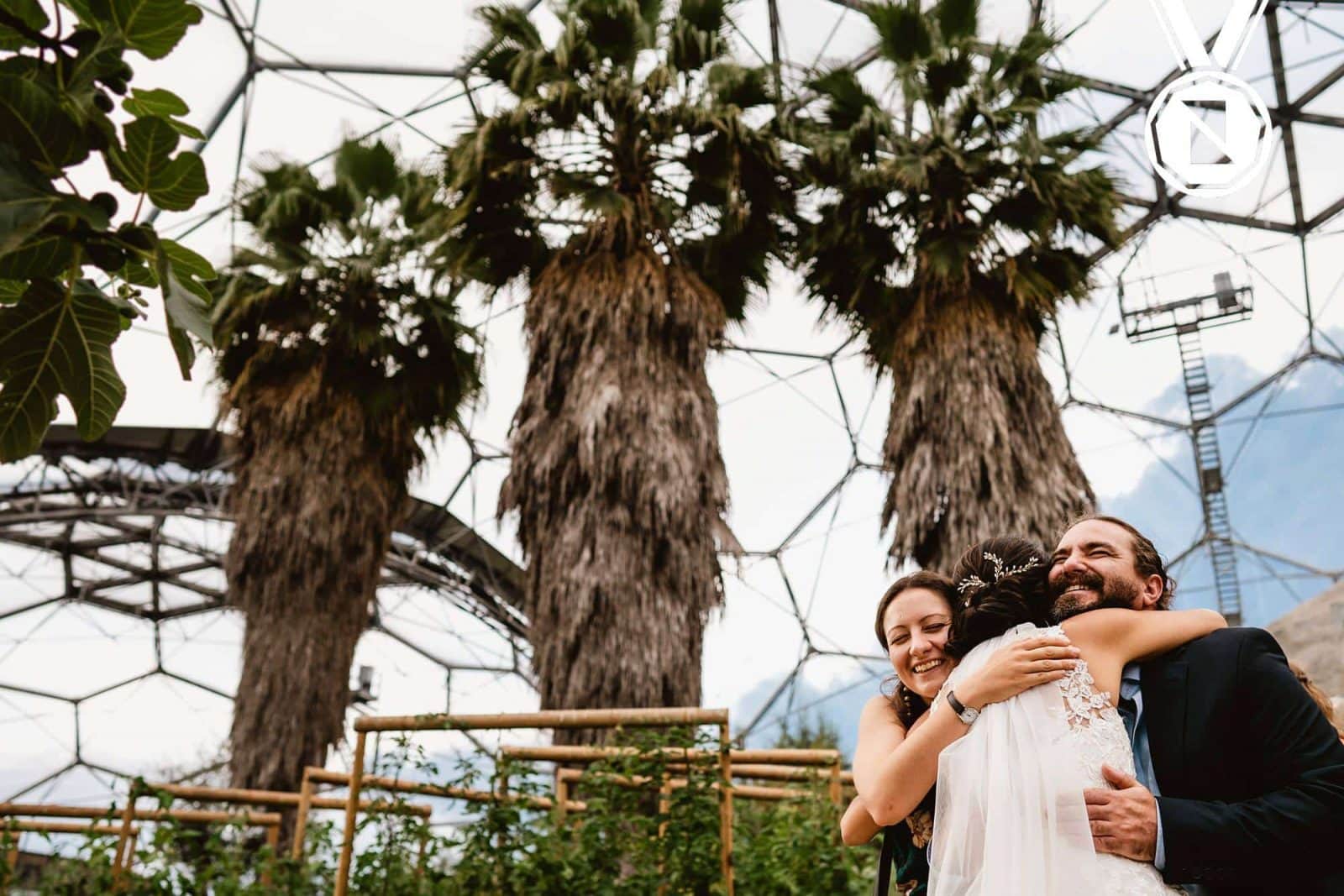 A wedding image at the Eden Project of a bride and two guests embracing infront of some huge trees.